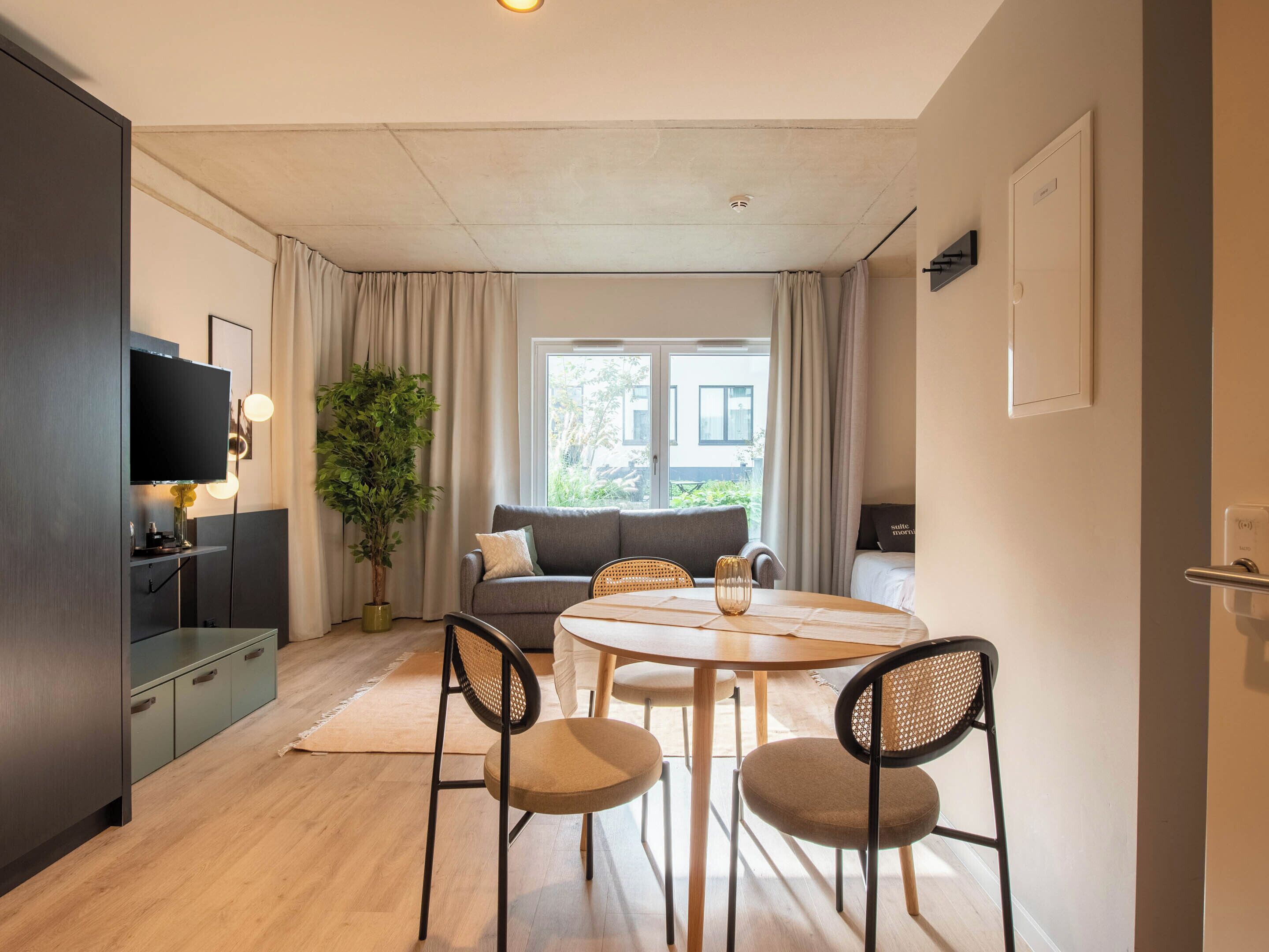 A modern living room with a sofa, a dining table with chairs, a television, and a large window with curtains.
