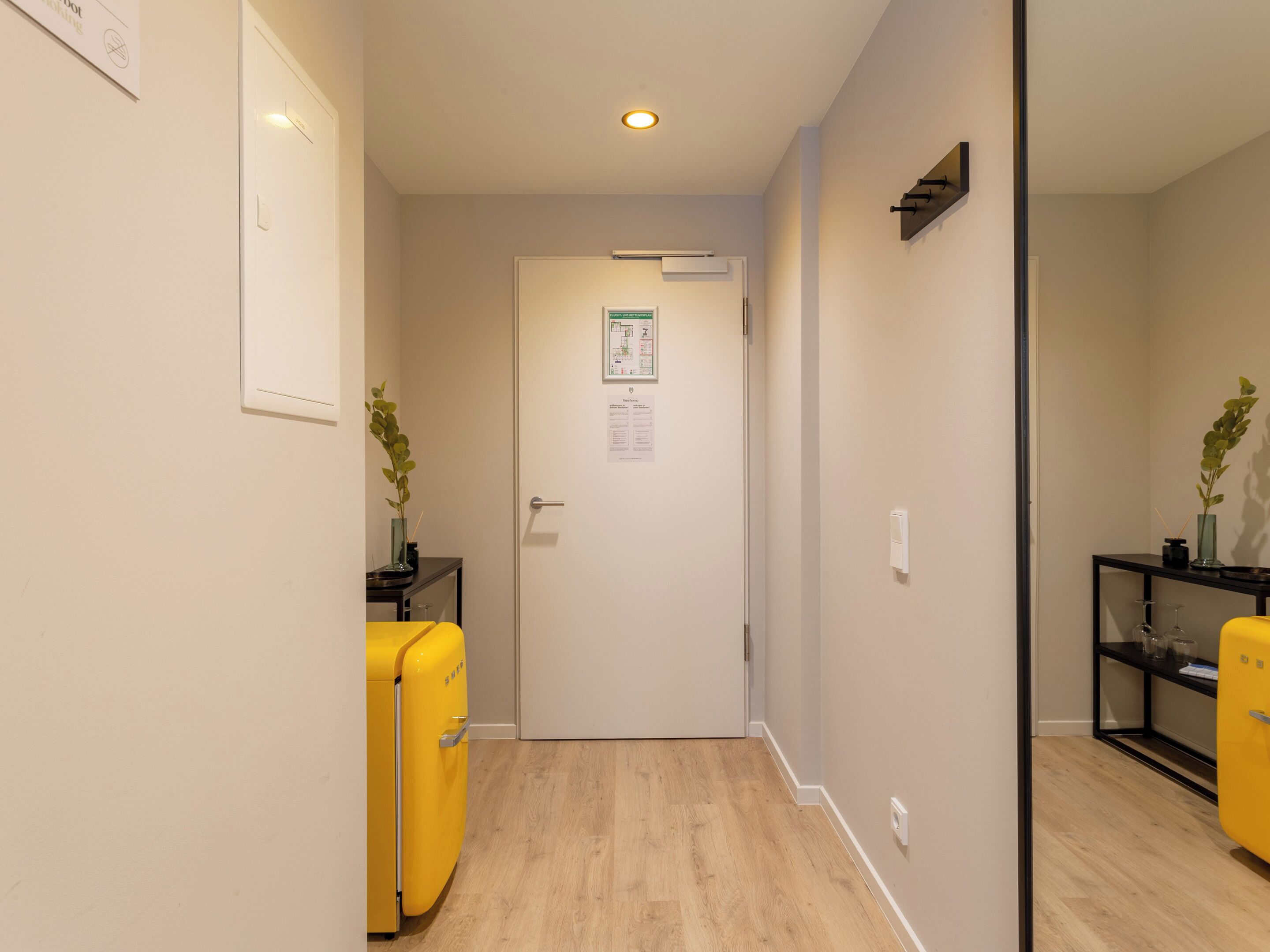 Un pasillo estrecho con piso de madera, una puerta con un letrero verde y un refrigerador amarillo a la izquierda.