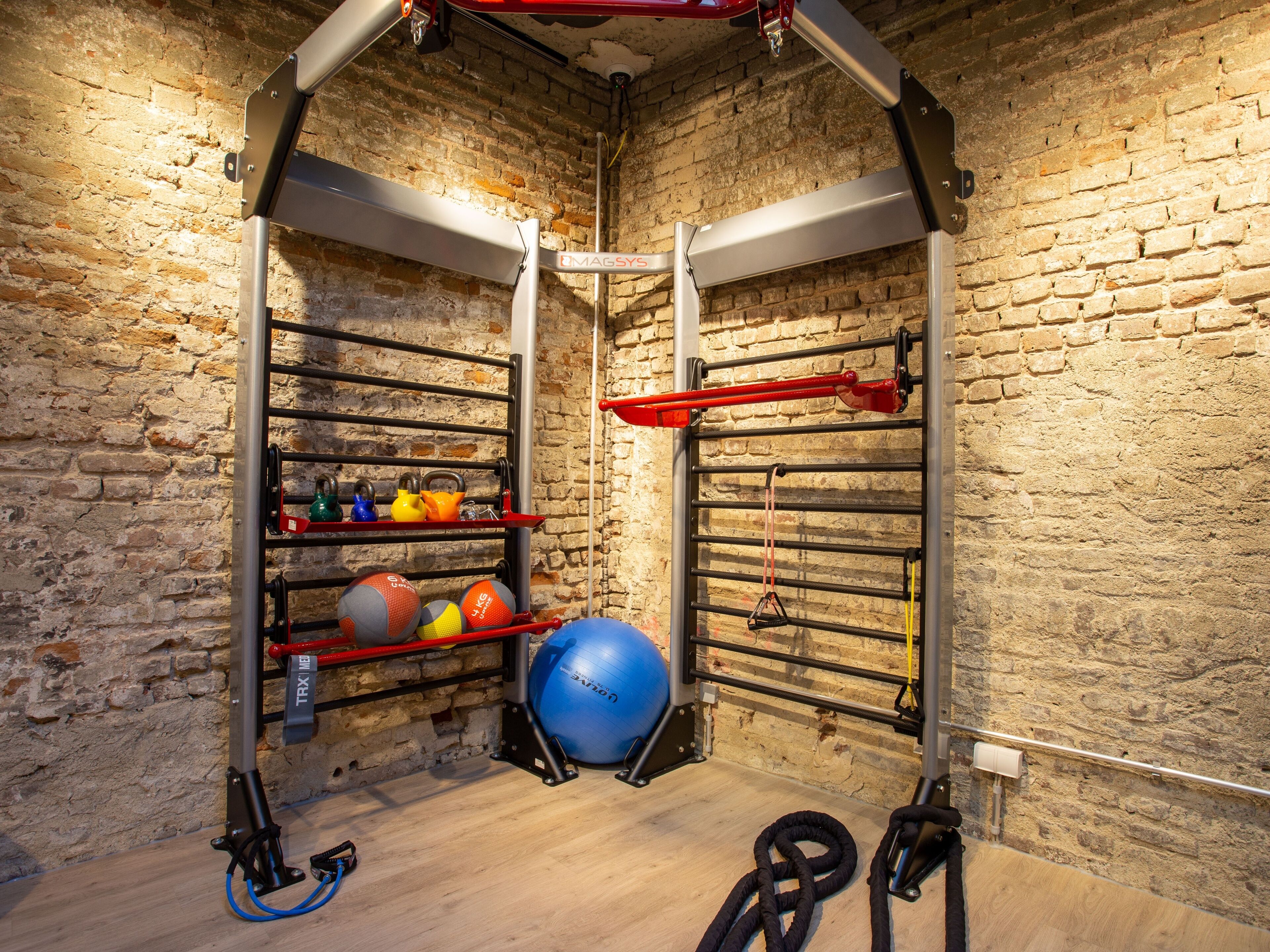 Ein Fitnessraum mit Backsteinwand, Metallregalen und Trainingsgeräten wie Kettlebells und Medizinbällen.