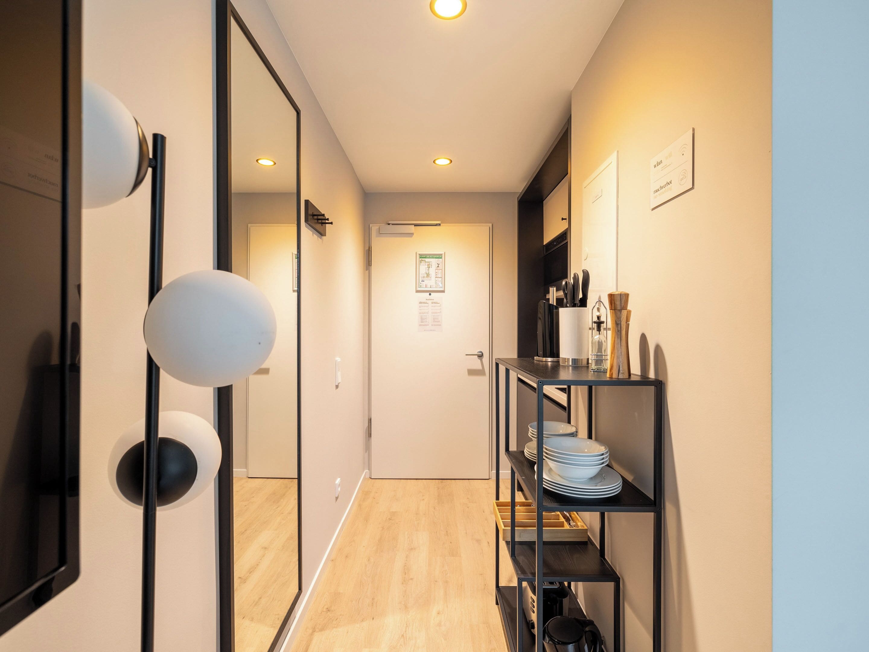 Un pasillo moderno de hotel con suelo de madera, puerta de cristal y repisas con utensilios de cocina.