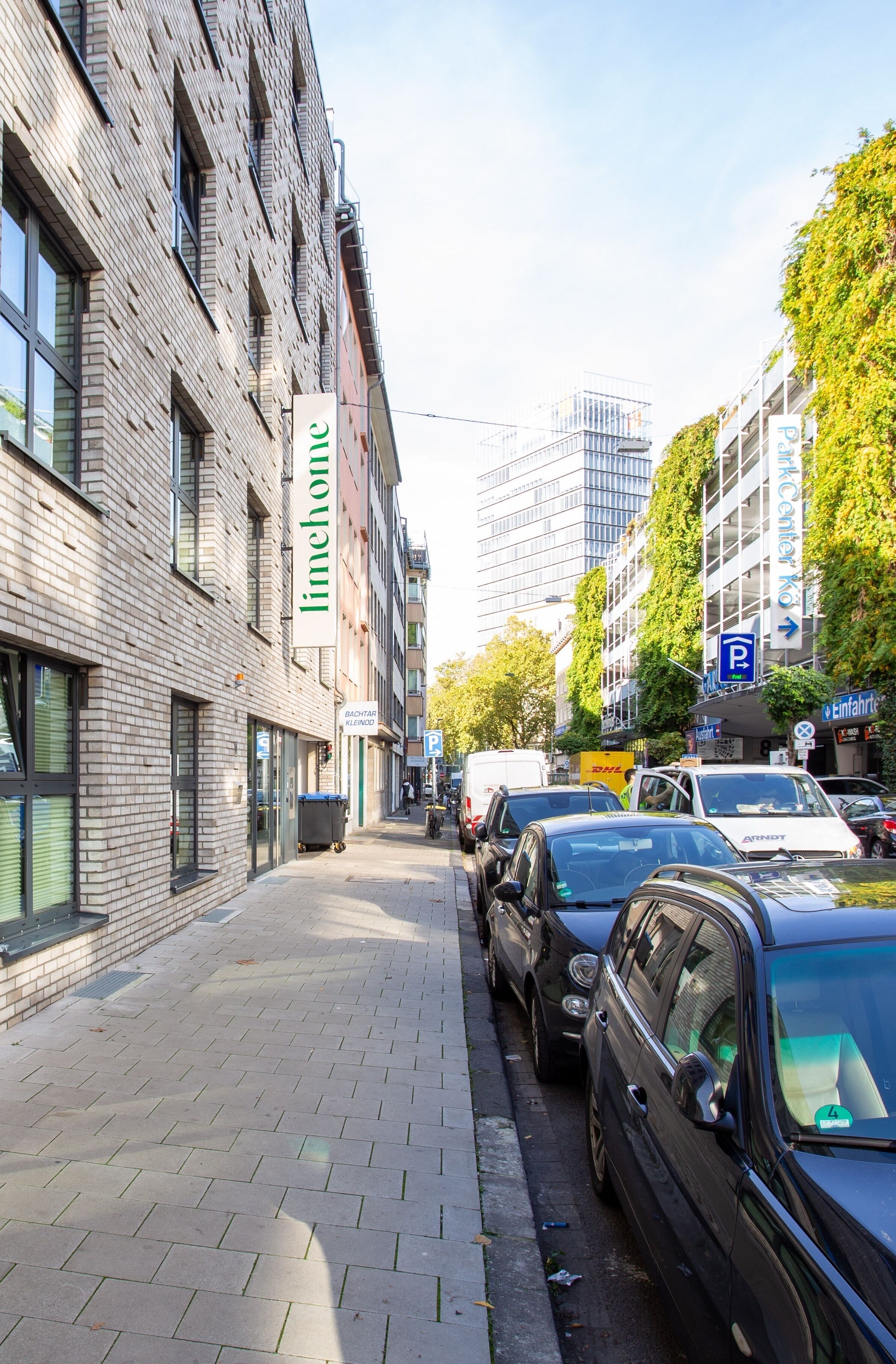 Une rue bordée de voitures garées, un immeuble avec une enseigne indiquant « imhouse », et un panneau de signalisation indiquant un parking.