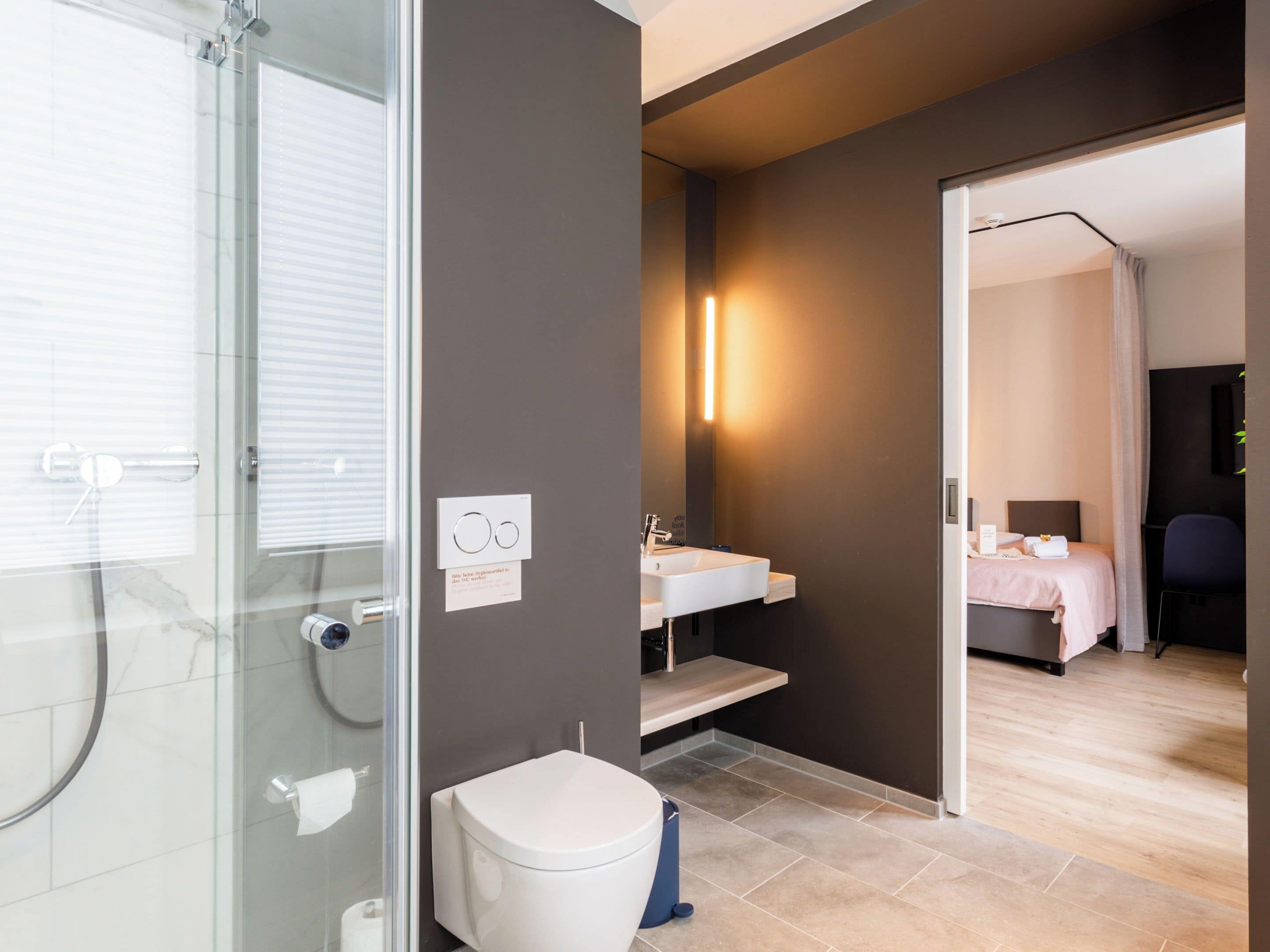 Un bagno moderno con box doccia in vetro, un WC bianco, un lavandino con specchio e un comodino con un letto nella stanza adiacente.