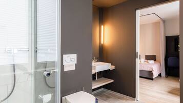 Un bagno moderno con box doccia in vetro, un WC bianco, un lavandino con specchio e un comodino con un letto nella stanza adiacente.