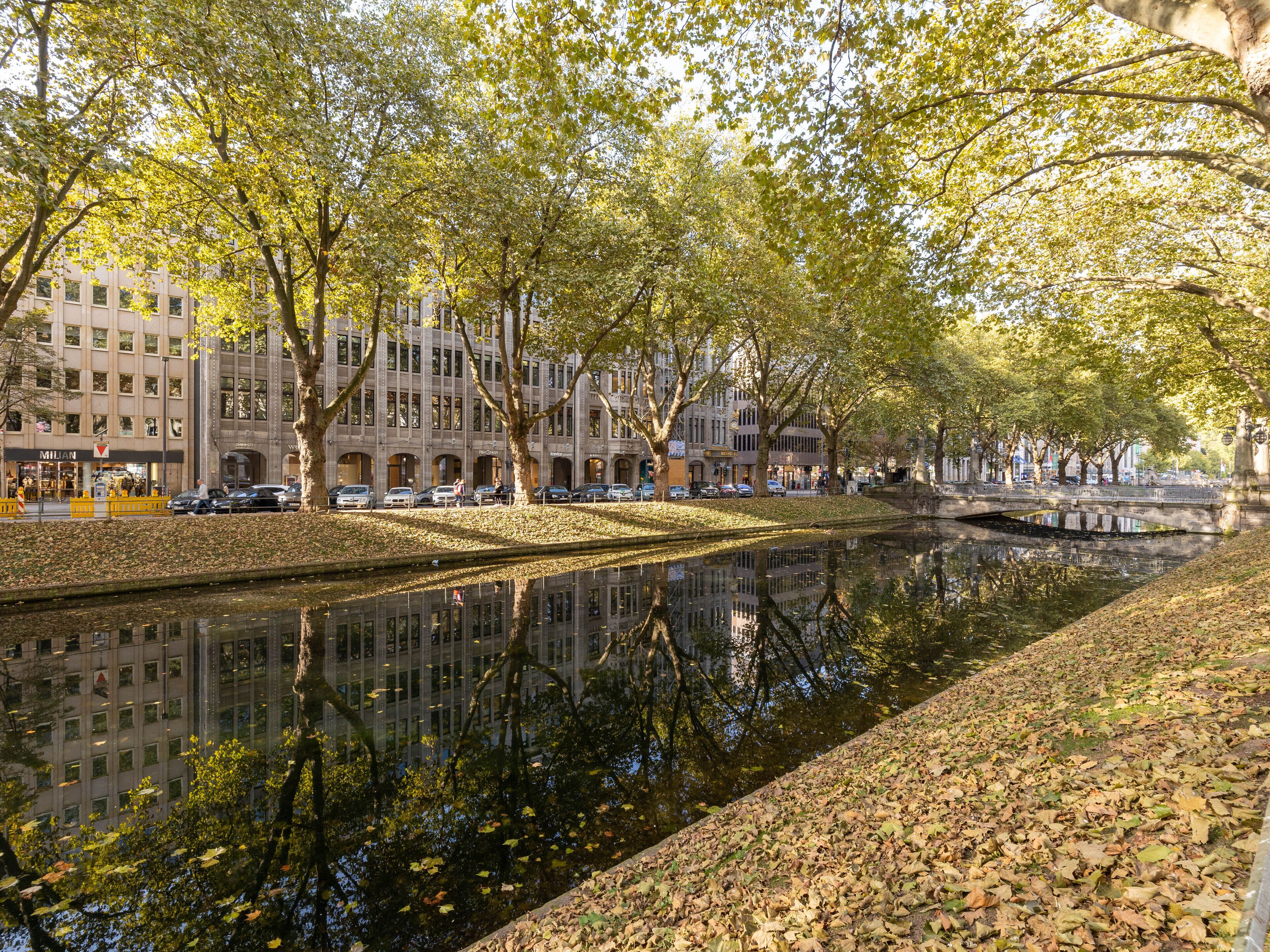A canal with trees reflecting in the water, a bridge, and a building with multiple windows and a sign that reads "MILAN".