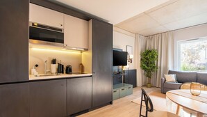 Een moderne keuken met donkere kasten, een ingebouwde oven en een aanrecht met een koffiezetapparaat. Er is een eethoek met een ronde tafel en stoelen, een bank en een groot raam met gordijnen.