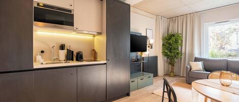 A modern kitchen with dark cabinetry, a built-in oven, and a countertop with a coffee maker. There is a dining area with a round table and chairs, a sofa, and a large window with curtains.