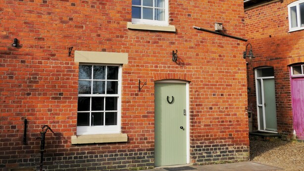 A red brick building with a green door, a window with a white frame, and a small birdhouse on the wall.