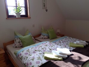 Un dormitorio con dos camas, almohadas verdes, colchas con motivos florales, una ventana con una planta y una lámpara fijada a la pared.
