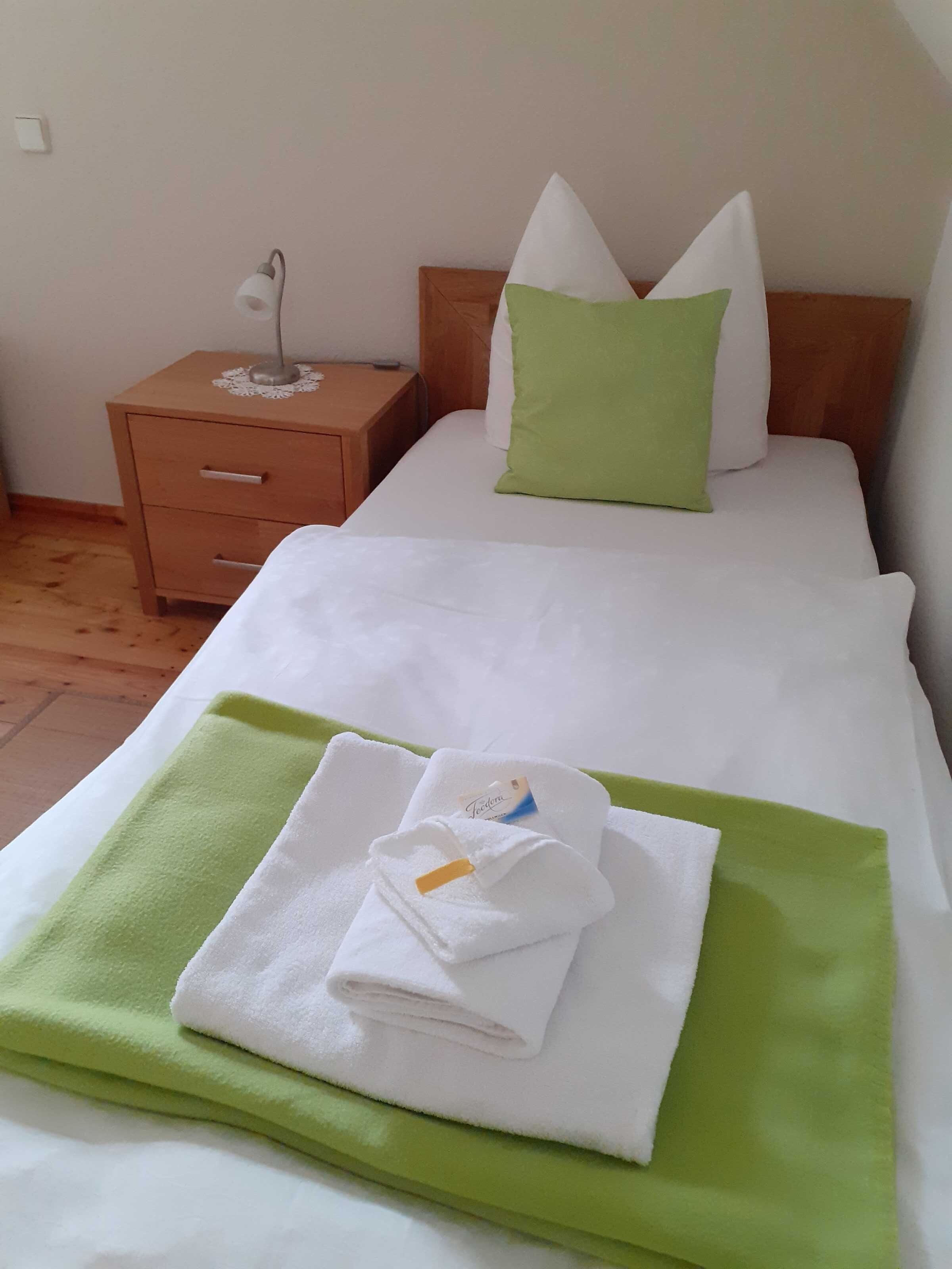 Un lit bien fait, avec une serviette verte et une serviette blanche pliée, une table de chevet en bois avec une lampe et une prise électrique au mur.