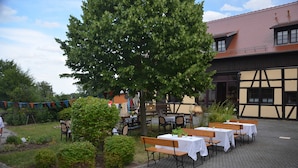 An outdoor dining area with tables and benches, surrounded by greenery and a building with a tiled roof.