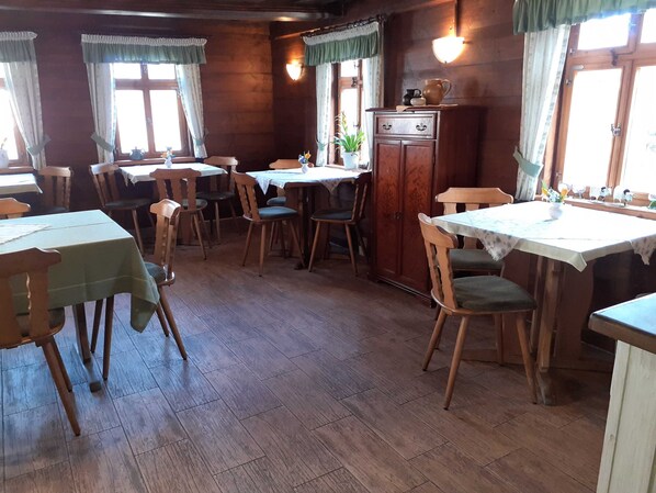 A dining area with wooden tables and chairs, a cabinet with decorative items, and windows with curtains.