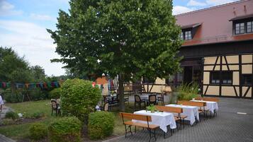 Un espace de restauration en plein air, avec des tables et des bancs, entouré de verdure et bordé d’un bâtiment dont le toit est recouvert de tuiles.