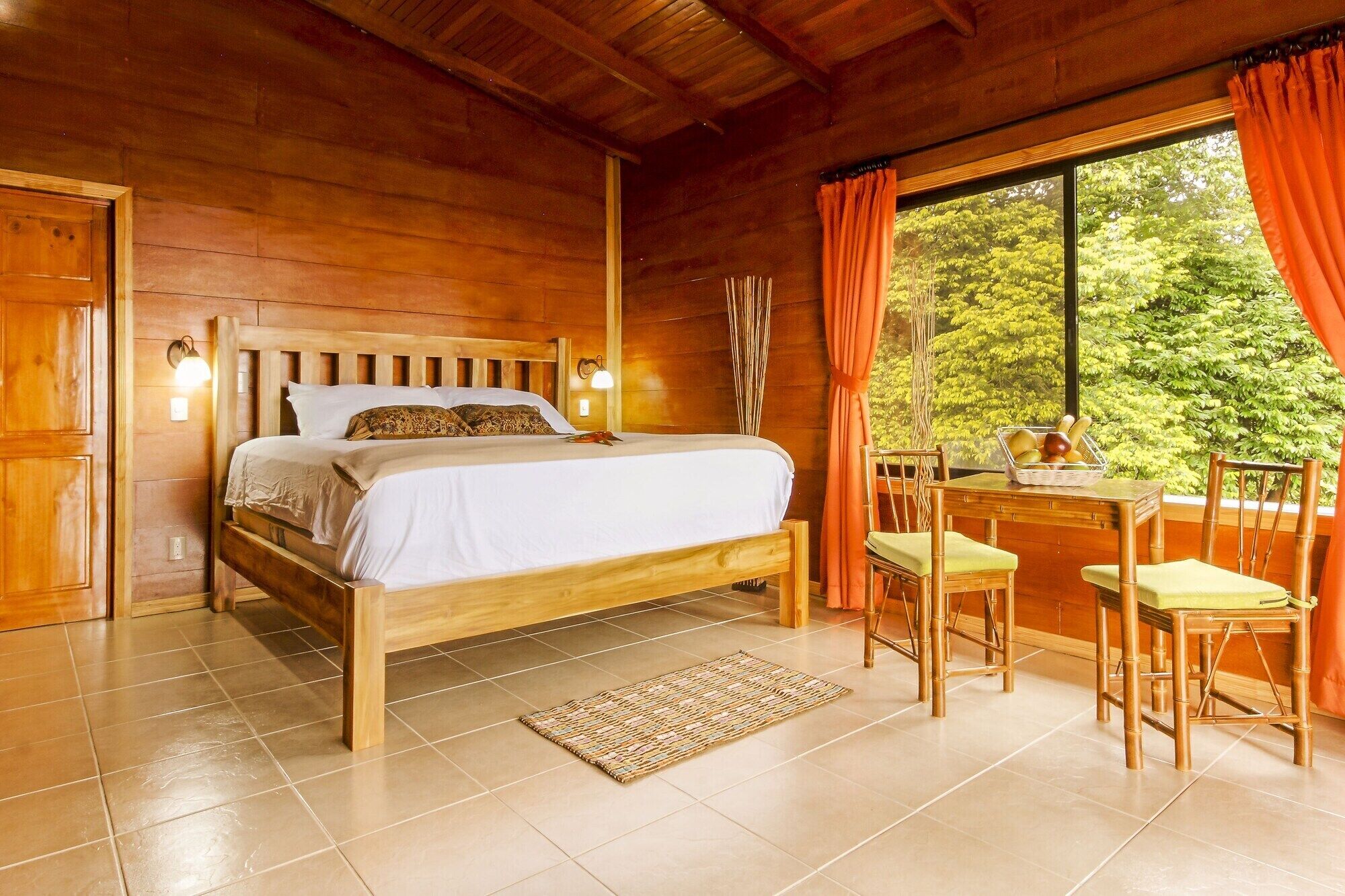 A wooden cabin bedroom with a bed, bedside lamps, a desk with chairs, and a view of greenery.