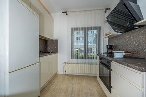 A modern kitchen with white cabinetry, a built-in oven, and a window with vertical blinds. - Apartment Dijana (Novi Vinodolski)