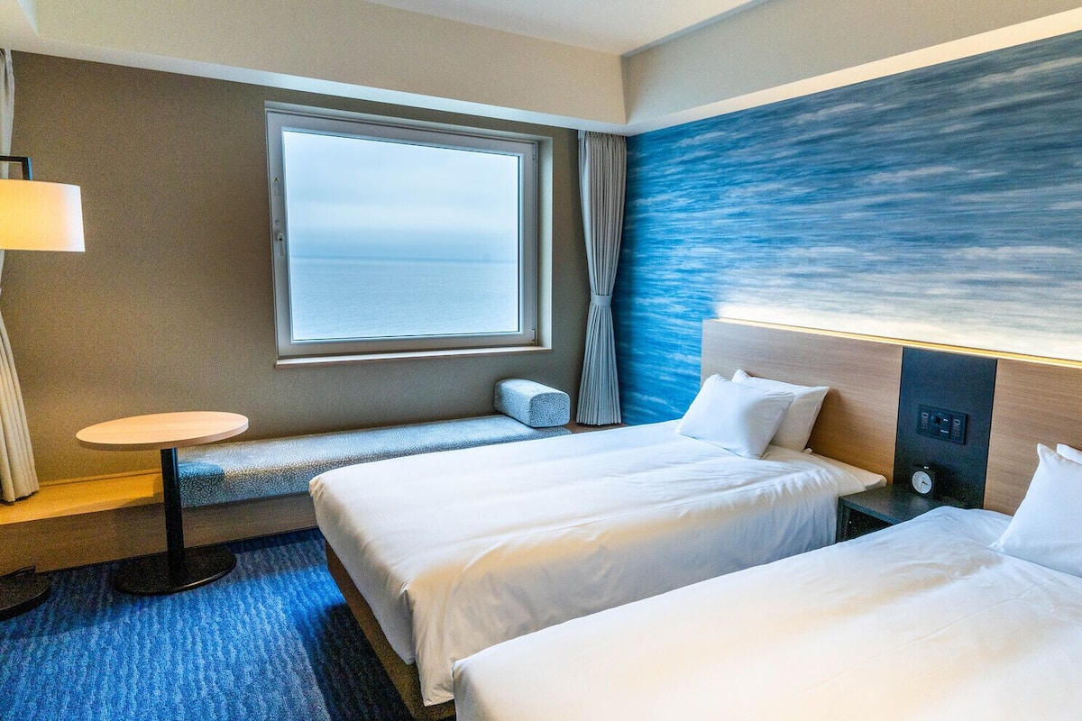 A hotel room with two beds, a small table, a lamp, a window with a view of the sea, and a wall with a blue abstract design.