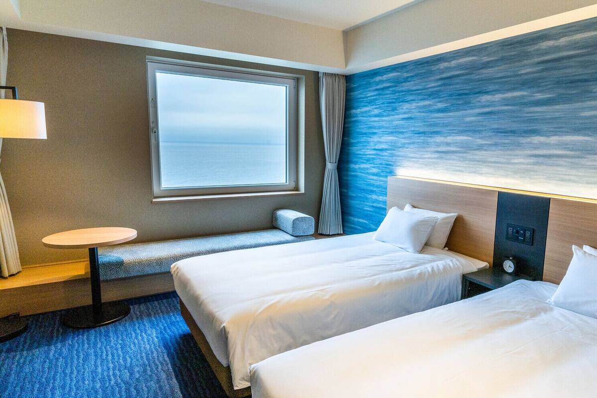 Ein Hotelzimmer mit zwei Betten, einem kleinen Tisch, einer Lampe, einem Fenster mit Meerblick und einer Wand mit blauem abstrakten Design.