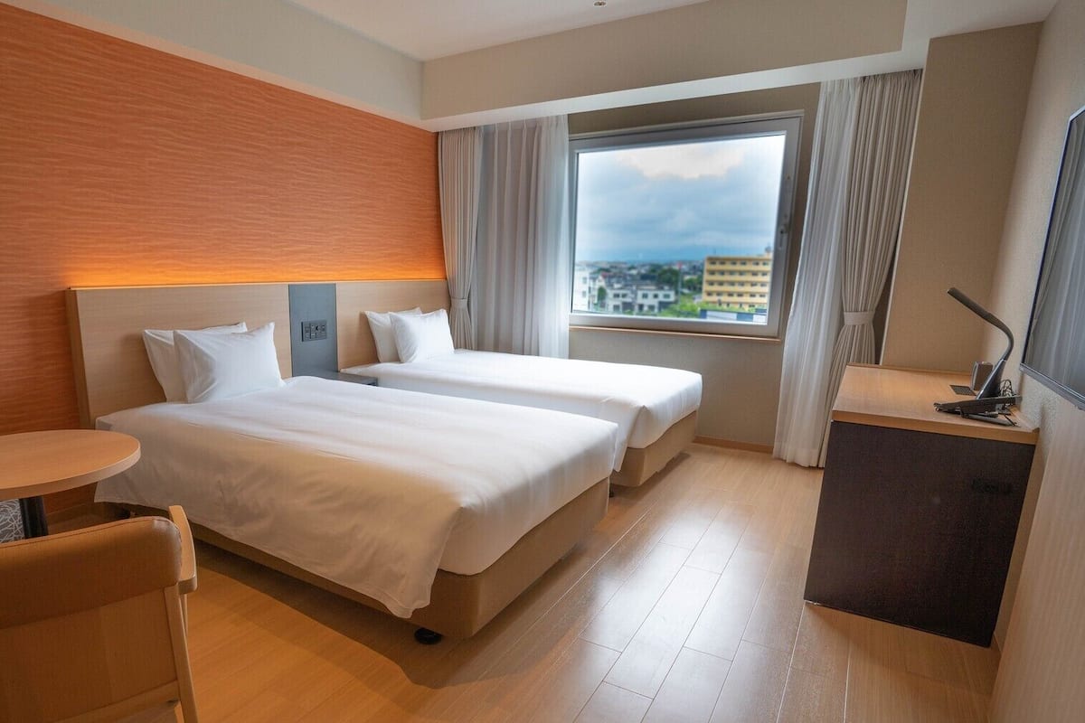 A hotel room with two beds, a small desk, a chair, and a view of the city.