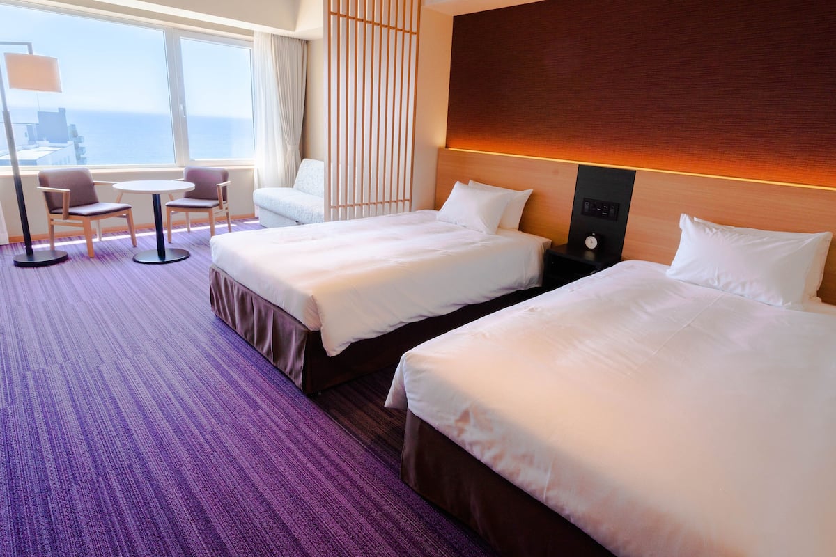 A hotel room with two beds, a small table, two chairs, a nightstand, and a view of the ocean.