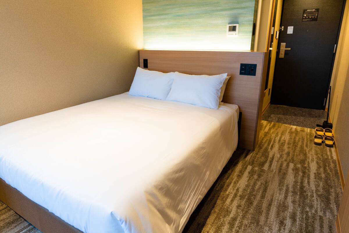 A hotel room with a bed, a headboard, a wall-mounted control panel, a door, and a carpeted floor.