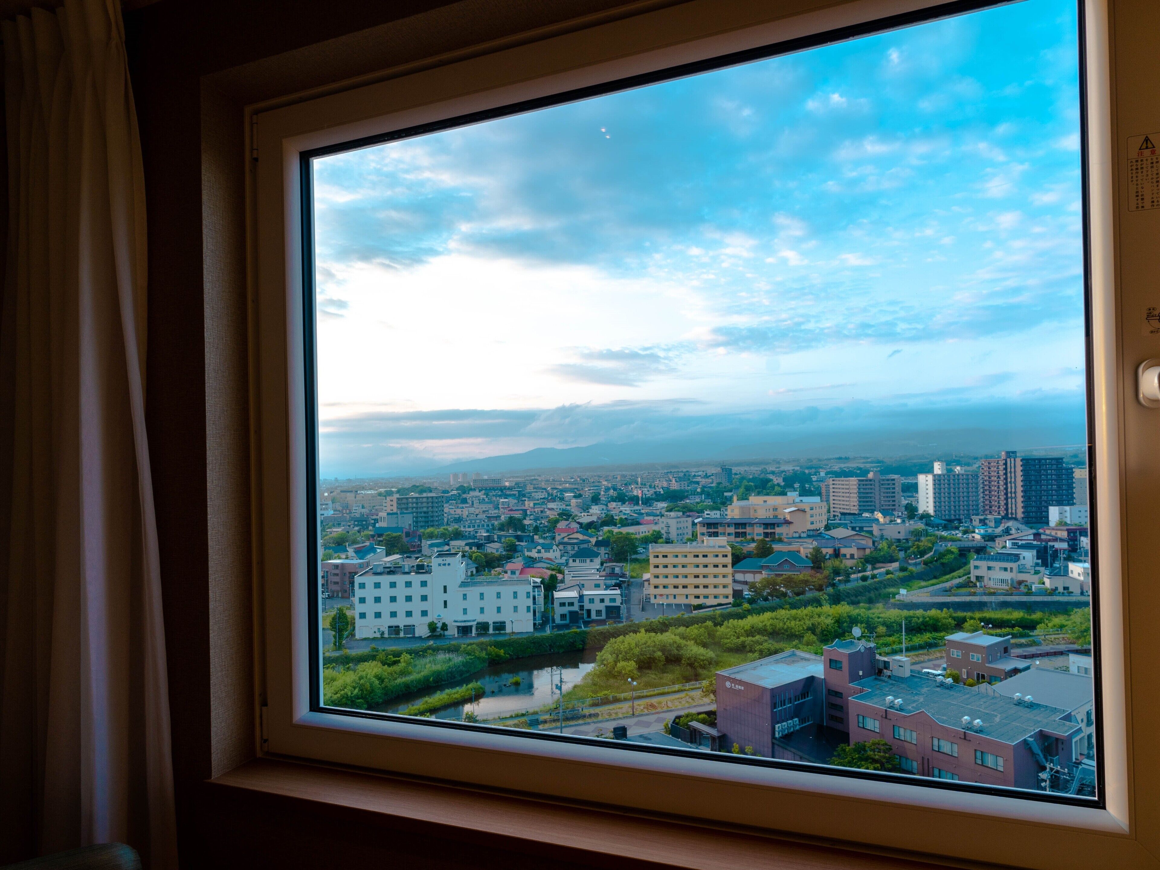 Vista desde una ventana que muestra un paisaje urbano con edificios, áreas verdes y una cadena montañosa distante.