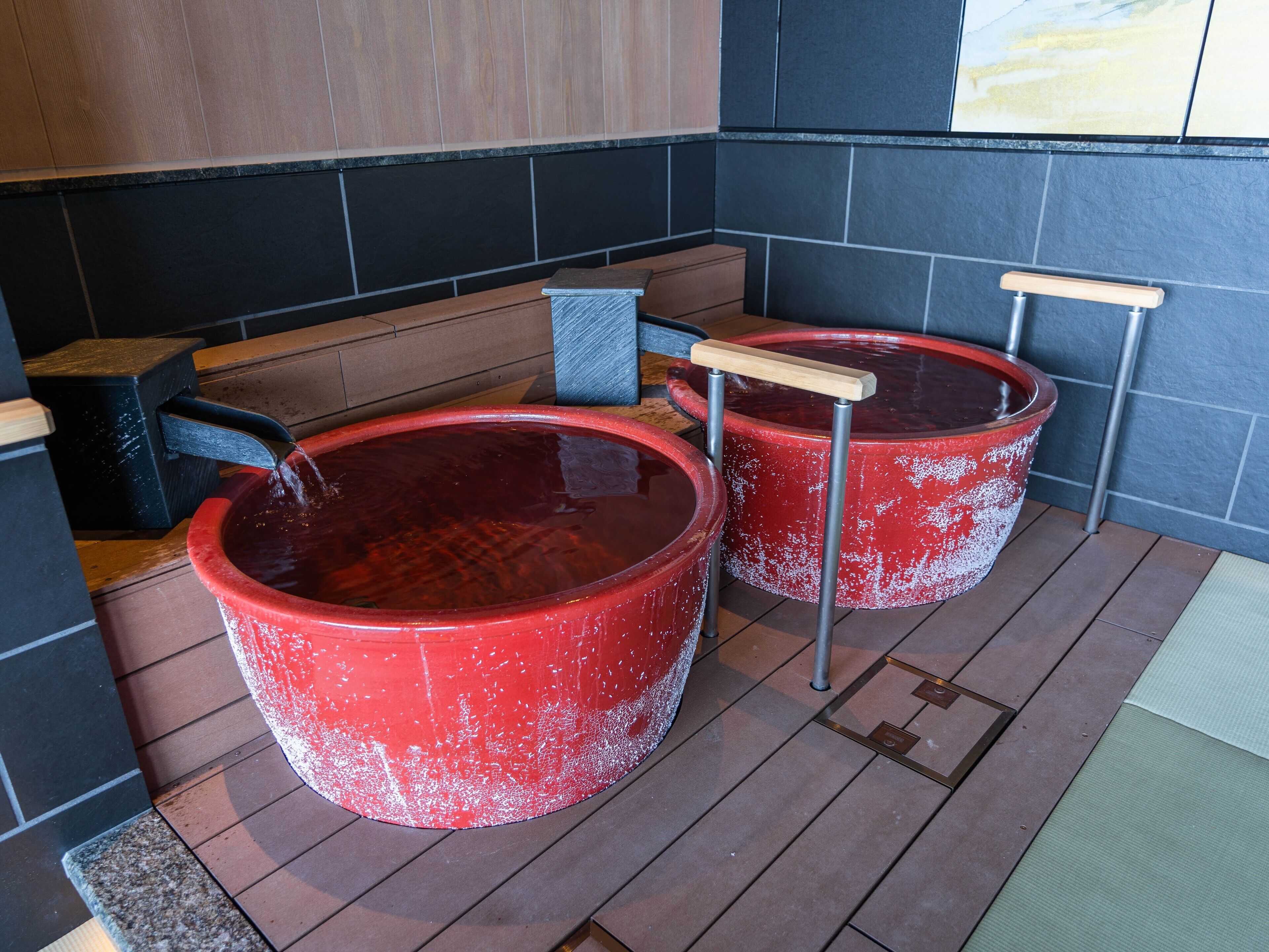 Two red hot tubs with wooden handles, set on a wooden deck with a tiled wall in the background.