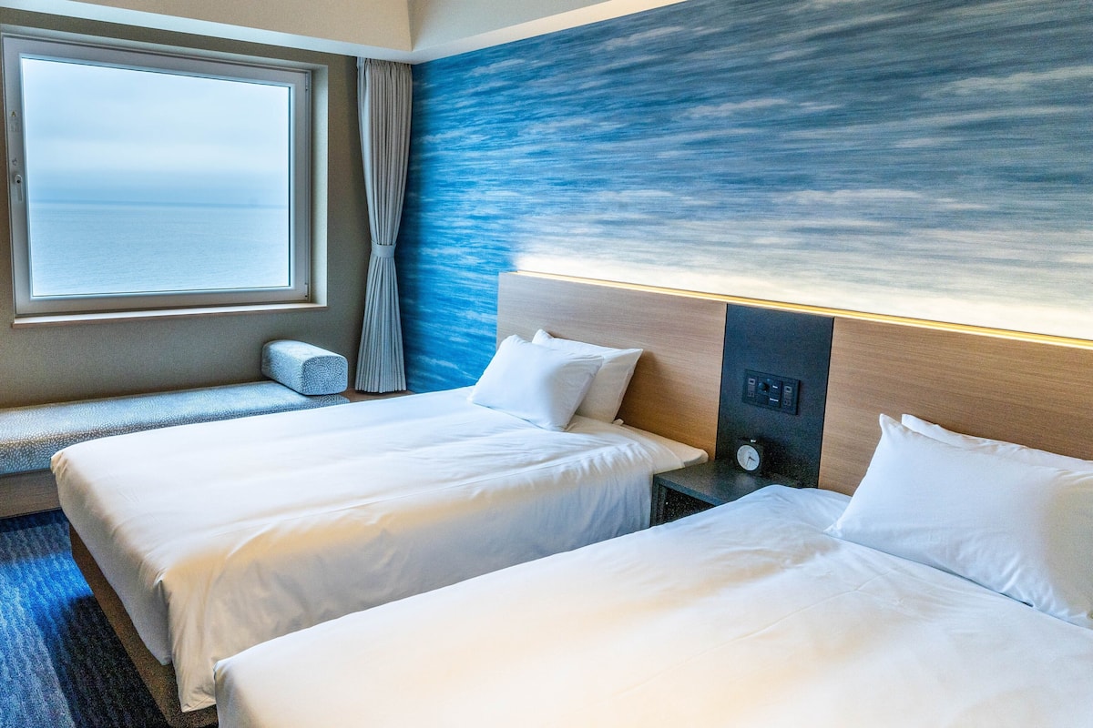 A hotel room with two beds, a large window with a view of the sea, and a blue textured wall.