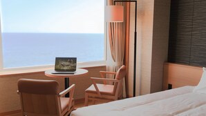 Une chambre d’hôtel avec un lit, deux chaises, une petite table ronde avec un ordinateur portable, une vue sur la mer et une grande fenêtre avec des rideaux.