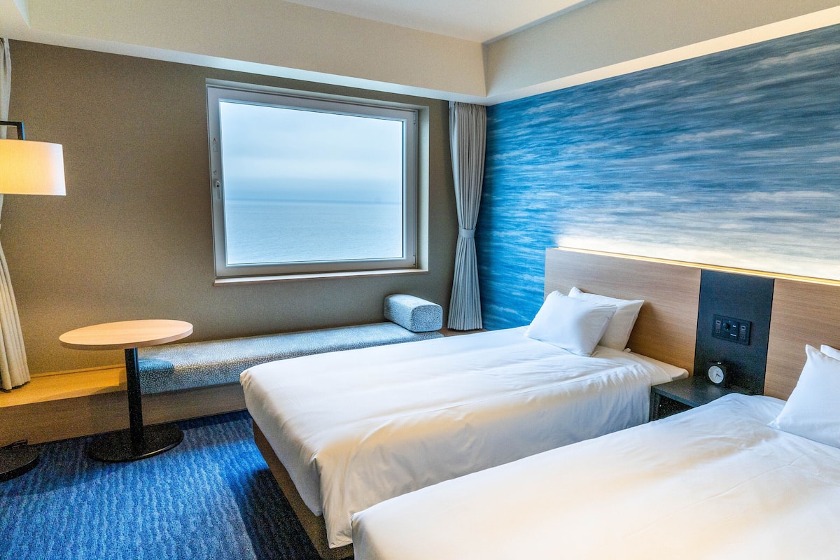 A hotel room with two beds, a small table, a bench, a window with a view, and a wall with a textured design.