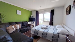 A hotel room with a green wall, a black leather sofa, a bed with patterned bedding, a small TV on a side table, and a view of the outdoors through a window. - Apartment Klara (Dramalj)