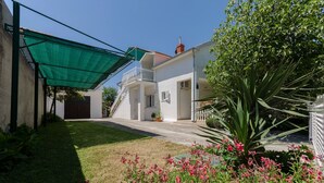 Un bâtiment blanc doté d’un balcon, d’un auvent vert et d’un jardin où poussent des fleurs roses.