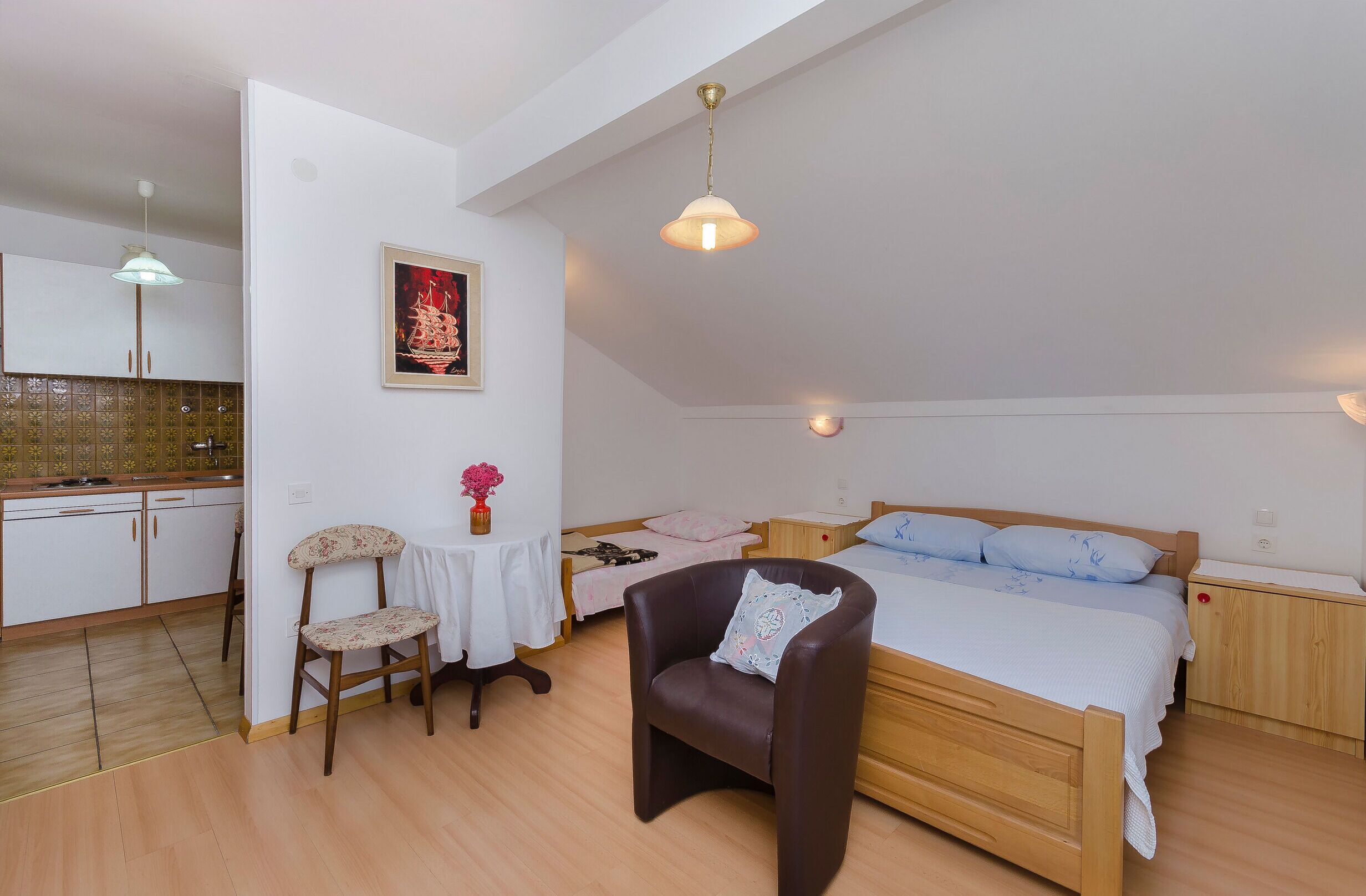 A bedroom with a wooden bed, a chair, a small table, and a kitchen area visible through an open door.