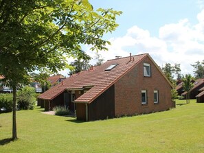 A single-story brick house with a sloped roof, a small attached wooden structure, and a well-maintained lawn.