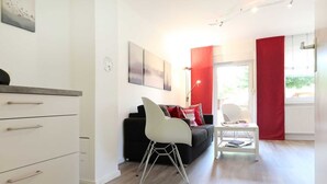 Een moderne woonkamer met een zwarte bank, een witte stoel en een kleine tafel. Er is een raam met rode gordijnen en uitzicht op groen.