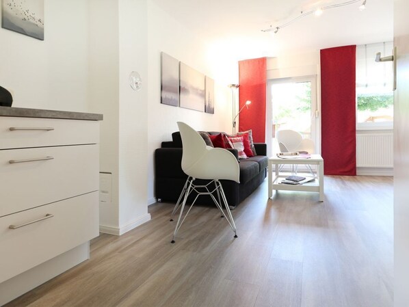 A modern living room with a sofa, a white chair, and a small table. There is a window with red curtains, a door, and a wall-mounted artwork.