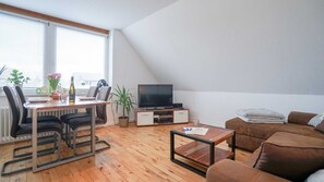 A modern living room with a dining area, a flat-screen TV on a wooden stand, a brown sofa, and a dining table with chairs.