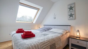 A bedroom with a sloped ceiling, a skylight, a bed with patterned bedding, red towels, a bedside table with a lantern, and a wall sign.