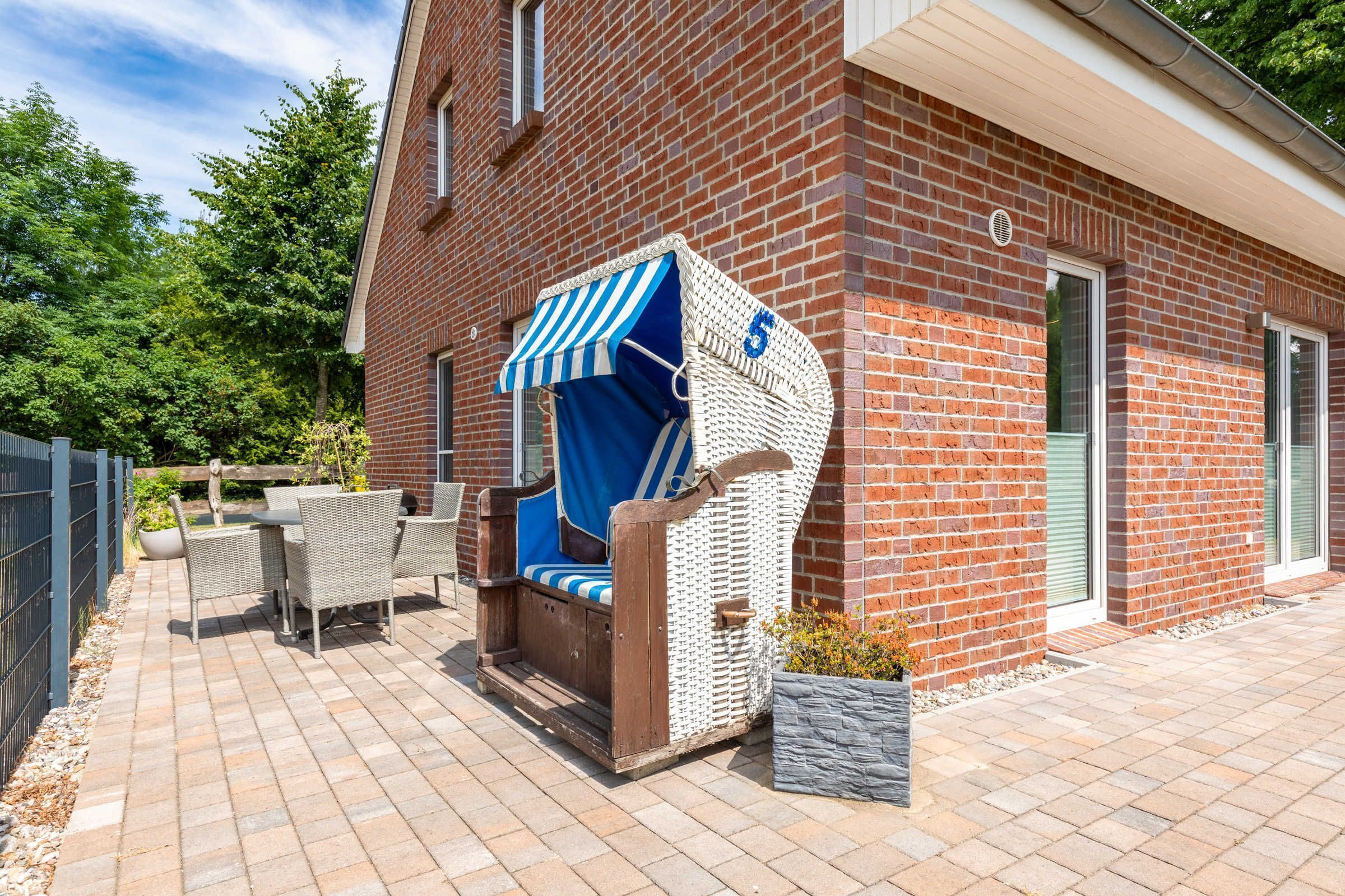 Ein Backsteinhaus mit einer Terrasse, ausgestattet mit Korbmöbeln, einem blau-weiß gestreiften Markisenvorzelt und einem Steinpflanzgefäß.