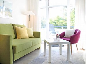 Quarto com um sofá verde, uma mesa branca, uma cadeira roxa e janelas grandes.