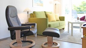 Un salon moderne avec un fauteuil en cuir, un canapé vert, un pouf en bois, une table blanche et un lampadaire.