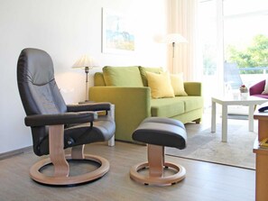 Un salon moderne avec un fauteuil en cuir, un canapé vert, un pouf en bois, une table blanche et un lampadaire.
