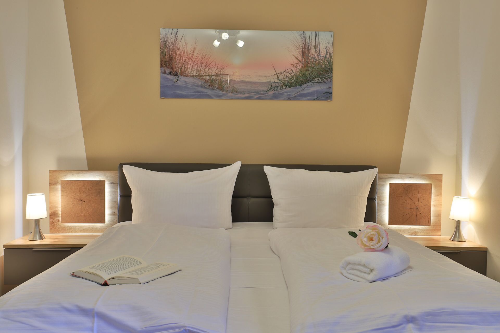 Una habitación de hotel con una cama grande, dos mesitas de noche con lámparas, un cuadro en la pared y un libro sobre la cama.