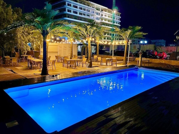 A swimming pool with lounge chairs and palm trees, surrounded by a building with multiple floors and balconies.