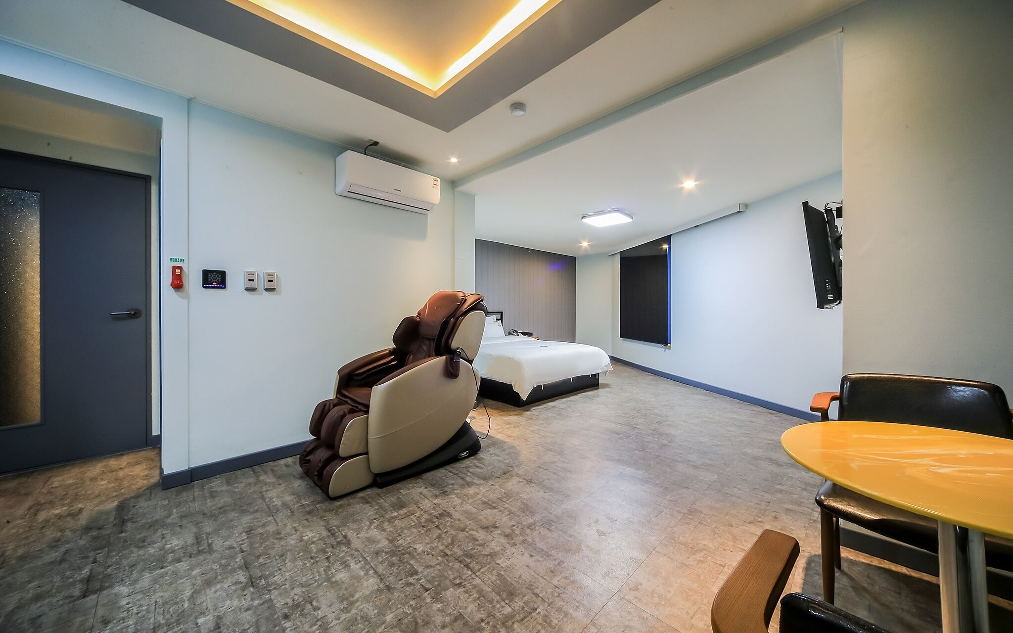 A hotel room with a massage chair, a bed, a small table, and a TV mounted on the wall.