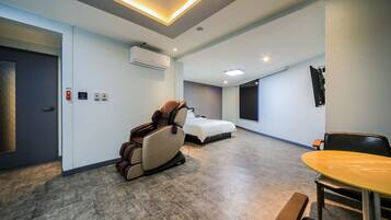 A hotel room with a massage chair, a bed, a small table, and a TV mounted on the wall.