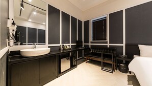 Une salle de bain moderne avec un grand miroir, un lavabo blanc, un meuble-vasque noir, un banc en cuir noir et un lit avec une literie blanche.