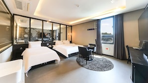 A hotel room with two beds, a desk, a chair, a round rug, and a large window with curtains.