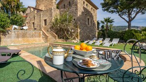 A poolside table with a teapot, a bowl of oranges, and pastries, set against a backdrop of a stone building with a tower. - Can Xirau Castle | Private Pool Sea Views Air Conditioning Ideal Families (SANTA SUSSANA)