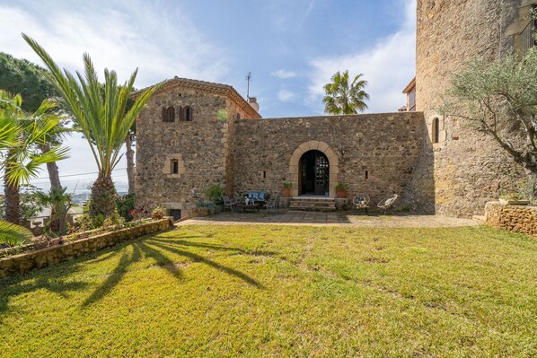 Un edificio de piedra con un arco central, un pequeño porche con sillas y un área verde al frente.