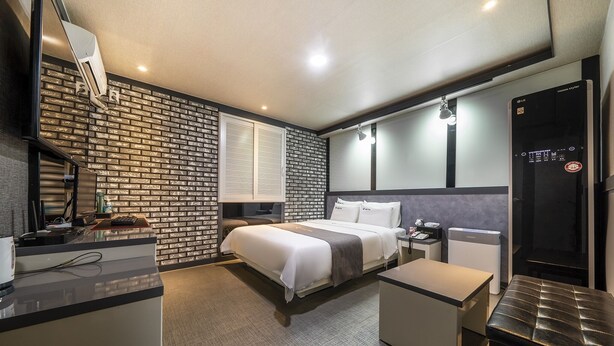 A modern hotel room with a brick-patterned accent wall, a bed with white linens, a desk with a computer, a small table, and a minibar. - Cheonan E (Cheonan)