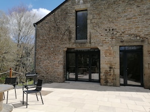 A stone building with a large glass door and windows, outdoor seating with chairs and a table, and a paved patio area. - Grande Propriété le Long du Ruisseau (ST NOLFF)